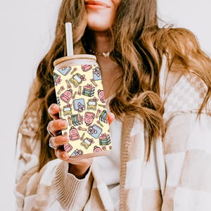 May include: A glass tumbler with a bamboo lid and straw. It has a repeating pattern of gift boxes, backpacks, and pencils in pastel colours on a yellow background. The cylindrical tumbler appears to contain a clear liquid.