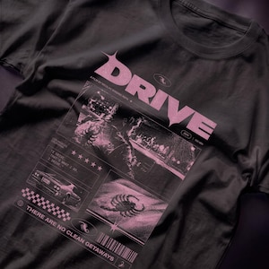 Può includere: Una t-shirt grigio antracite con una stampa grafica rosa che riporta la parola DRIVE, un fotogramma di un film, un'auto, uno scorpione e il testo: A FILM BY NICOLAS WINDING REFN, 2011, 100 MIN, LOS ANGELES CALIFORNIA, I DRIVE... THAT'S WHAT I DO. I DRIVE, THERE ARE NO CLEAN GETAWAYS.