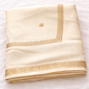 May include: A folded cream-colored fabric featuring a wide, intricate gold embroidered border along the bottom edge and a thinner, matching gold embroidered border forming a corner frame, with a small, circular gold embroidered motif in the center.