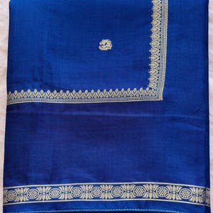 May include: A folded, royal blue textile with gold-colored decorative borders. The borders feature floral and geometric patterns. A small, gold-colored floral design is in the upper left corner. The fabric appears to be silk or a similar material.