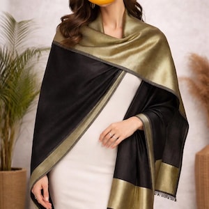 May include: A black and gold shawl with a metallic sheen. The shawl has a wide gold border and a contrasting black body. The fabric appears soft and drapes elegantly. The shawl is worn over a white dress, creating a sophisticated look.