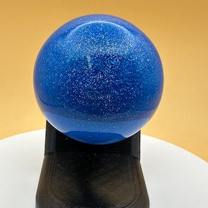 May include: A blue sphere with glitter suspended within, resting on a black, textured stand. The sphere has a glossy finish and gradients of blue. The stand is a solid, geometric shape.