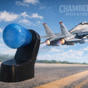 May include: A blue, bulbous object on a black stand, with the text "CHAMBERLIN SPEED & CUSTOM" in the background. A fighter jet is taking off in the background against a blue sky.