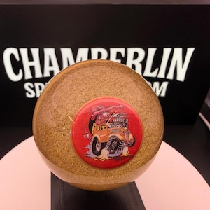 May include: A gold glitter-covered sphere with a red button featuring a cartoon hot rod. The sphere is set against a black background with the white text "CHAMBERLIN SP". The object is likely a decorative item.