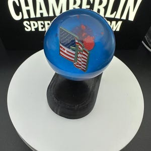 May include: A blue, spherical object with an American flag and cross design. The object is mounted on a black base and sits on a white circular platform. The word "CHAMBERLIN" is visible in the background.