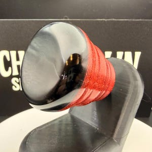 May include: A black and red object with a shiny, rounded top and a textured red section. The object is mounted on a black, angular base. The background features the text "CH S".