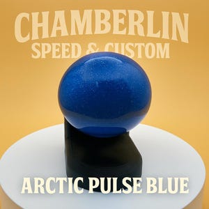 May include: A blue sphere with a speckled appearance, labeled "Arctic Pulse Blue," rests on a black stand. The background is a warm yellow-orange, with the text "Chamberlin Speed & Custom" above the sphere.