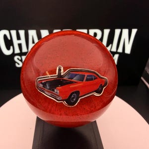 May include: A red, spherical shift knob with a car emblem. The knob has a speckled texture and a shiny finish. The car emblem is a red muscle car with a black hood. The background has the text "CHAMBERLIN".
