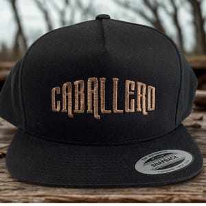 Könnte beinhalten: Schwarze Snapback-Kappe mit dem Wort "CABALLERO" in beige auf der Vorderseite gestickt. Die Kappe hat einen flachen Schirm und einen Druckknopfverschluss hinten. Ein kleines silbernes Etikett befindet sich auf dem Schirm.