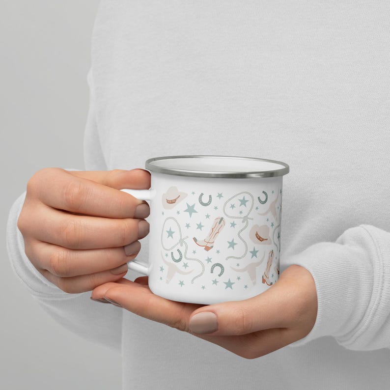 May include: White enamel mug with a silver rim, featuring a repeating pattern of cowboy hats, boots, horseshoes, and stars in light blue, beige, and gray. The mug is held in a person's hands.