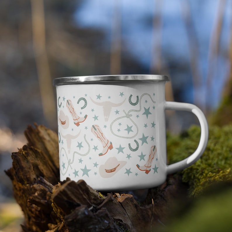 May include: White enamel mug with a silver rim featuring a repeating pattern of cowboy boots, hats, stars, horseshoes, and longhorn skulls in muted colors. The mug has a white handle and is set against a blurred natural background.