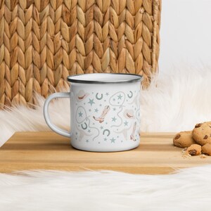May include: White enamel mug with a black rim, featuring a repeating pattern of cowboy hats, boots, horseshoes, and stars in light brown and blue-green hues. The mug sits on a wooden surface with cookies nearby.