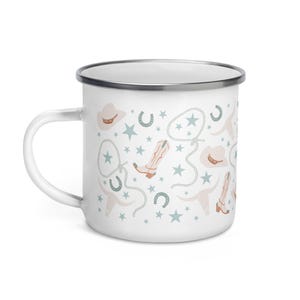 May include: White enamel mug with a cowboy-themed pattern. The design includes cowboy boots, hats, horseshoes, stars, and rope in shades of beige, light blue, and brown. The mug has a silver rim and a white handle.