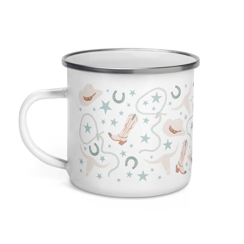 May include: White enamel mug with a cowboy-themed pattern. The design includes cowboy boots, hats, horseshoes, stars, and rope in shades of beige, light blue, and brown. The mug has a silver rim and a white handle.