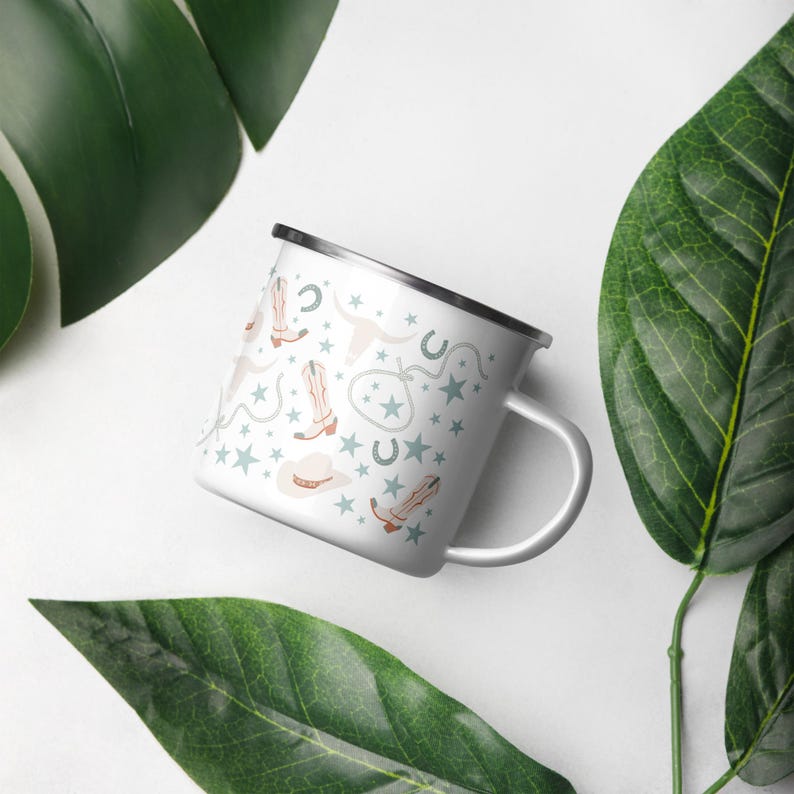 May include: White enamel mug with a silver rim and handle. The mug features a repeating pattern of cowboy boots, hats, stars, horseshoes, and steer heads in light brown and blue. The mug is set against a white background with green leaves.