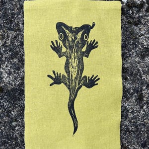 May include: A rectangular, light yellow fabric with a black lizard print. The lizard is stylized with a long tail and outstretched limbs. The fabric has a textured appearance, and the lizard design is centered on the fabric.
