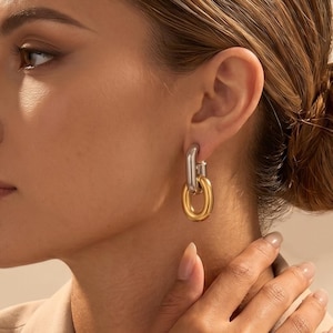 Könnte beinhalten: Zweifarbige Gold- und Silber-Creolen. Die Ohrringe haben ein Doppelschleifen-Design, mit einem silbernen Oberteil und einer goldenen unteren Schlaufe. Die Ohrringe werden von einer Person getragen, die einen beigen Blazer trägt.