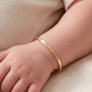 Könnte beinhalten: Ein goldfarbenes Babyarmband mit der Aufschrift "EMMA MY LOVE" ist am Handgelenk eines Babys zu sehen. Das Armband hat ein schlichtes Banddesign. Das Baby trägt einen cremefarbenen Strampler.