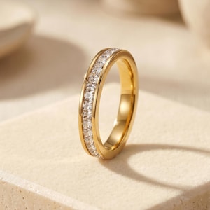 May include: A gold-colored ring with a band of clear, square-cut stones. The ring is set on a light beige surface, with soft lighting and a blurred background. The ring is a piece of jewelry.