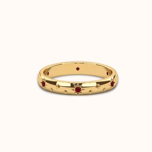 May include: A gold ring with a band featuring a repeating pattern of stars and red gemstones. The ring has a polished finish and is set against a white background. The gemstones are evenly spaced around the ring.