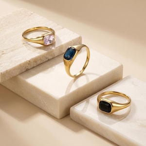 Peut inclure: Trois bagues en or avec des pierres précieuses sont présentées sur une surface texturée blanc cassé. Une bague présente une pierre rose clair, une autre une pierre bleu foncé et la troisième une pierre noire. Les bagues ont un design classique et élégant.
