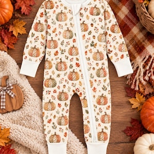 May include: A long-sleeve baby romper featuring a cream background with an all-over print of various pumpkins, autumn leaves, and acorns. The garment includes a full-length front zipper closure and solid white ribbed cuffs at the wrists and ankles.