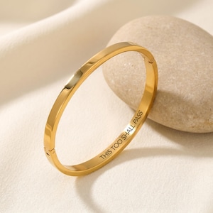 Könnte beinhalten: Ein goldfarbenes Armreif-Armband mit dem eingravierten Text "THIS TOO SHALL PASS". Das Armband hat ein schlichtes, elegantes Design und einen Verschluss. Das Armband wird auf einer weißen Oberfläche neben einem glatten, hellfarbigen Stein präsentiert.