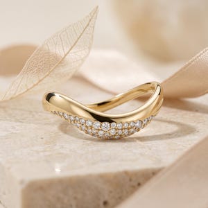 May include: A curved gold ring featuring a polished band with a pavé setting of numerous small, clear, round-cut stones along the front section. The metal has a smooth, reflective finish, and the design follows a gentle, undulating wave shape.