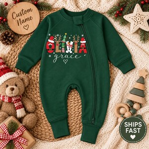 Op de afbeelding: Een donkergroene baby-romper met lange mouwen, een diagonale ritssluiting en een feestelijke afbeelding op de borst. De afbeelding toont de naam Olivia Grace met letters gestileerd als de Kerstman, rendieren, een kerstboom en een sneeuwpop.