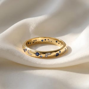 May include: A gold ring with a celestial design. The ring features blue and clear gemstones in a pattern of stars, moons, and dots. The inscription "S925" is visible. The ring is set against a white, textured background. A ring of this type would be measured in centimetres.