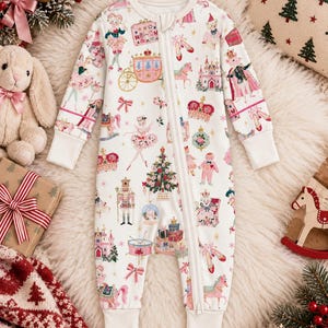 May include: A white long-sleeve baby romper featuring a full-length front zipper and a colorful print of Nutcracker-themed illustrations, including ballerinas, crowns, castles, gift boxes, rocking horses, and Christmas trees with pink and gold accents.