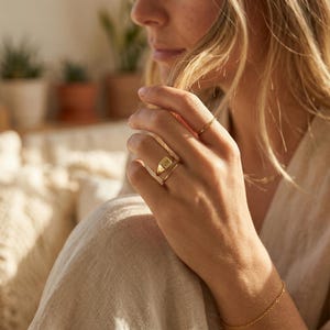 Könnte beinhalten: Ein goldener Siegelring mit den Initialen "QM" auf der Vorderseite. Der Ring wird an einem Finger getragen, zusammen mit einem dünnen goldenen Bandring. Auch ein zartes goldenes Armband ist zu sehen.