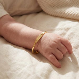 Op de afbeelding: Een goudkleurige babyarmband met de inscriptie "MY TINY TREASURE" is te zien om de pols van een baby. De armband is gebogen en gepolijst. De hand van de baby rust op een crèmekleurige stof.