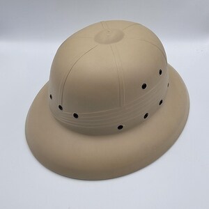 Puede incluir: Sombrero safari de color beige con ala ancha y copa redondeada. El sombrero presenta una serie de pequeños orificios de ventilación negros espaciados uniformemente alrededor de la copa. Líneas verticales recorren los lados.