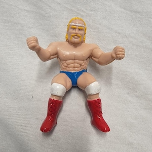 May include: A vintage action figure of a muscular wrestler with blonde hair and a mustache. The figure is wearing blue trunks, red boots, and white knee pads. The wrestler is posed with arms raised.