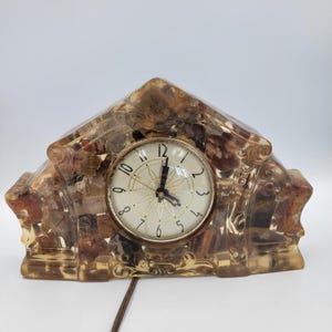 May include: Vintage mantel clock with a clear, amber-colored resin case. The clock face is white with gold-colored hands and numerals. The clock has a decorative, ornate design and a brown power cord.