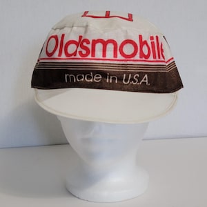 Puede incluir: Gorra Oldsmobile blanca y marrón con visera curva. La gorra presenta el logotipo de Oldsmobile en rojo y el texto "made in U.S.A." en marrón. La gorra se muestra en una cabeza de maniquí blanca.