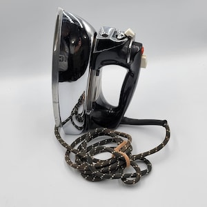 May include: Vintage electric iron with a chrome and black finish. The iron has a curved design with a handle and control buttons. The power cord is brown and white, coiled at the base. A classic appliance.