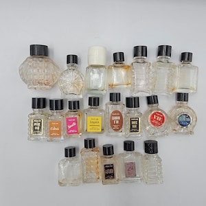 May include: A collection of vintage perfume bottles in various shapes and sizes. The bottles are mostly clear glass with black or white caps. Some bottles have labels with French text, including "Parfum Magic Rose" and "Croyance".