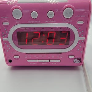 Puede incluir: Reloj despertador digital rosa con una pantalla LED roja que muestra la hora. Cuenta con botones para programar, detener, saltar y reproducir. Incluye una pantalla de CD y función de reposo. El reloj tiene un gráfico de Hello Kitty.