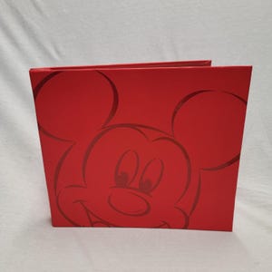 Puede incluir: Un álbum de fotos rojo con una imagen sutil en relieve de Mickey Mouse. El álbum, de forma cuadrada, tiene un acabado mate. El diseño es un tema clásico de Disney, perfecto para guardar recuerdos.