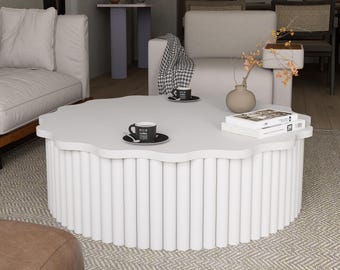 May include: A white, scalloped-edge coffee table with a cylindrical base. Two black coffee cups and saucers sit on the table, along with a stack of books. The table is in a living room setting with a neutral color palette.