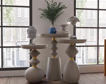 May include: Three decorative side tables with unique geometric designs. The tables are primarily gray with yellow accents and are topped with various decorative items, including a vase with greenery and a Buddha head sculpture. The tables are placed in front of a large window.