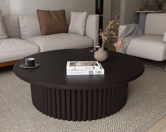 May include: A dark brown, round coffee table with a cylindrical base. The table has a smooth top and is approximately 40 inches in diameter. Two books and a cup and saucer are on the table. The table is in a living room setting.