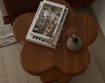 May include: A wooden, flower-shaped coffee table with a dark brown finish. Two magazines and a small gray vase with dried flowers rest on the table. The magazines have black and white images and text.