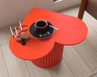 May include: A vibrant red heart-shaped side table with a cylindrical base. A black coffee cup and saucer with a spoon sit on top. Two candle holders with candles are also on the table. The table is placed on a light wood floor.