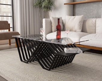 May include: A modern coffee table with a clear glass top and a black, geometric base. The table has a lower shelf and is in a living room setting with a white sofa, wine glasses, and a bottle of red wine.
