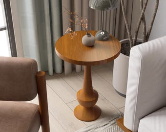 May include: A round, wooden side table with a light brown finish. The table has a pedestal base with a bulbous design and a circular top. A small vase with dried flowers and a green lamp sit on the table.