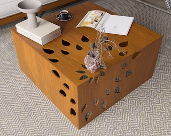 May include: A square, wooden coffee table with a warm brown finish. The table features leaf-shaped cutouts on the sides and top. A stack of books, a decorative vase, and a cup of coffee sit on the table.