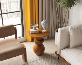 May include: A small, round wooden side table with a stacked, spherical base. The tabletop holds a small bowl, a stack of books, and a white bust sculpture. The table is in a room with a window, curtains, and a sofa.